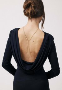 Femme aux cheveux relevés en chignon, portant une robe noire à manches longues avec un dos ouvert drapé et profond, et un collier chaîne en or tombant dans le dos.