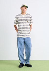 Stripet kortermet skjorte i kremfarge og svart, med en brystlomme og logo. Kombinert med avslappede blå jeans og svart fottøy. Grønn gulv.