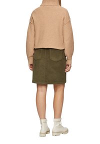 Korte beige gebreide trui met geribde kraag, gecombineerd met een olijfgroene corduroy rok met zakken. Witte enkellaarsjes maken de look compleet.