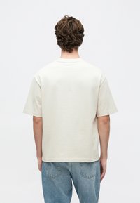 Młoda osoba z falującymi brązowymi włosami, ubrana w gładką jasnobeżową koszulkę i jasnoniebieskie dżinsy, stojąca tyłem na białym tle.