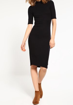 Femme portant une robe noire ajustée longueur genou avec manches courtes et des bottines marron, debout devant un fond blanc uni.