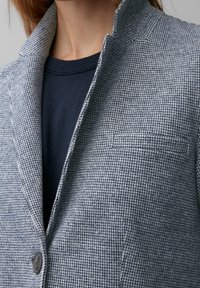 Blazer s houndstooth vzorem v šedé a námořnické modré, s klopovým límcem, uzavíráním na jeden knoflík a mírným polstrováním ramenních částí.