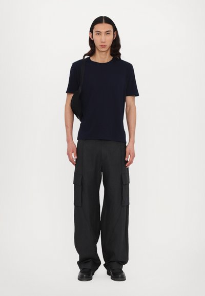 Marineblauw T-shirt, relaxte pasvorm; zwarte cargobroek met zijzakken; zwarte schoenen; het model staat tegen een effen witte achtergrond.