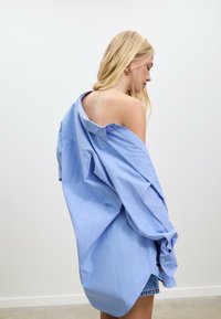 En blond kvinde iført en oversized, lyseblå stribet skjorte, der hænger ned fra den ene skulder og afslører en ryg-tatovering, står mod en ensfarvet lys baggrund.