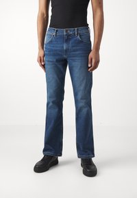 Jeans din denim albastru cu un design drept, având cinci buzunare și închidere cu nasture, asortați cu adidași negri. Textură netedă.