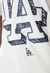 T-shirt en coton blanc avec un grand motif bleu marine et des lettres en contour. En bas, un petit logo de marque avec "LA" est affiché.
