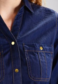 Camicia di jeans di un blu scuro con bottoni a pressione dorati, tasche sul petto e cuciture a contrasto. Presenta un colletto classico e una texture morbida.