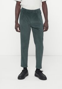 Groene corduroy broek met een ontspannen pasvorm, voorzien van een tailleband en subtiele verticale ribbels. Gepaard met zwarte schoenen.