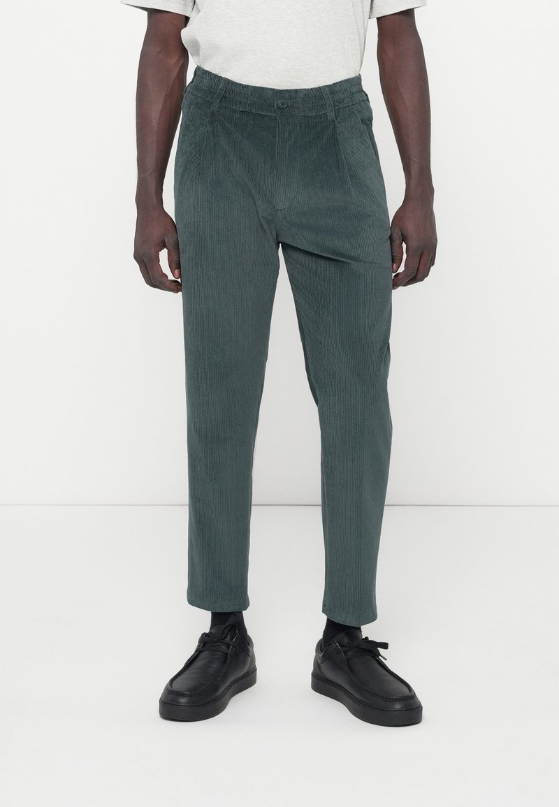 Groene corduroy broek met een ontspannen pasvorm, voorzien van een tailleband en subtiele verticale ribbels. Gepaard met zwarte schoenen.