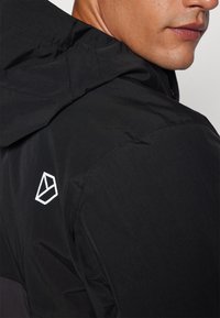 Black hooded jacket made from a lightweight material. Features a visible white logo on the shoulder, with a smooth texture and minimalist design.