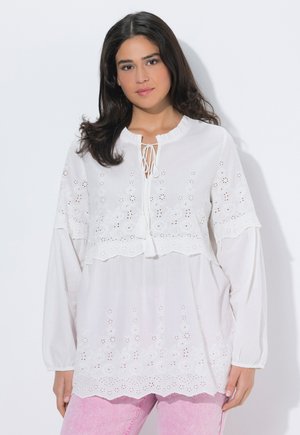 Kobieta o ciemnych włosach ubrana w białą haftowaną bluzkę z długim rękawem z wiązaniami na frędzle i jasnoróżowe spodnie na białym tle.