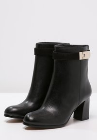 Botines de cuero negro con una textura suave, tacón grueso y una correa de ante en contraste con un detalle de hebilla metálica.