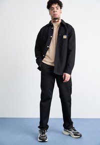 Sort skjorte med knapper over en beige t-shirt, sorte bukser og sorte og grå sneakers. Indeholder et lille logo på skjortens lomme.