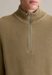 Brauner gestrickter Pullover mit hohem Kragen und Reißverschluss. Der Stoff hat ein strukturiertes Rippmuster und eine metallische Reißverschluss-Akzentuierung.