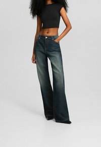 Kvinna klädd i mörkblå vida jeans och en svart kort topp, stående med en hand i fickan mot en enfärgad bakgrund.