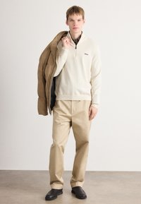 Pulover din tricot crem cu fermoar pe jumătate și logo, asociat cu o pereche de pantaloni bej larga și o jachetă de exterior marron. Pantofii negri tip slip-on completează look-ul.
