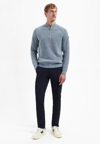 Lichtblauwe trui met een halve rits kraag en geribde manchetten, gecombineerd met marineblauwe broek en crèmekleurige sneakers, staande tegen een witte achtergrond.