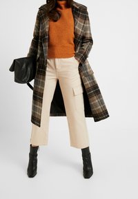 Długi płaszcz w kratę w ciemnozielonym i brązowym kolorze, pomarańczowy sweter z prążkowanym wzorem, beżowe spodnie typu cargo oraz czarne botki za kostkę. Charakteryzuje się wyraźną konstrukcją i fakturą.