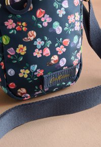 Marinblå crossbody-väska i tyg med mönster av flerfärgade blommor och snäckor samt en marinblå rem på en beige yta.
