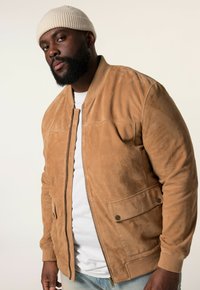 Tan Suede-Bomberjacke mit Reißverschluss, gerippten Bündchen und seitlichen Taschen, kombiniert mit einem weißen T-Shirt und einer hellbeigen Mütze.