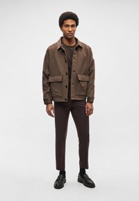 Casaco castanho texturado com grandes bolsos frontais, combinado com calças slim castanho-escuras e sapatos pretos. Modelo está em frente a um fundo neutro.