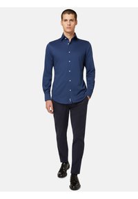 Mornarsko modra srajca z dolgimi rokavi in z gumbasto ovratnikom, opremljena z belimi gumbi, v kombinaciji s temnimi hlačami in črnimi vezalkami.