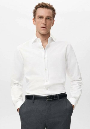 Camisa blanca de botones con cuello clásico y mangas largas, combinada con pantalones grises y un cinturón negro, con una textura de tela suave.