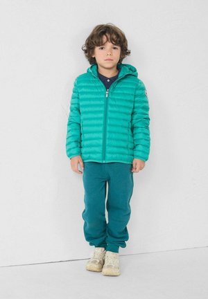Teal pufferjack met horizontale stiksels en een capuchon, gecombineerd met bijpassende teal joggingbroeken. Lichtgekleurde sneakers maken de outfit af.