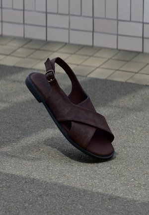 Sandalia de cuero marrón con diseño de tiras cruzadas, hebilla en el tobillo y suela de goma negra, sobre un fondo de pavimento texturizado.