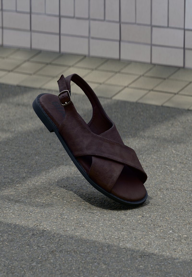 Καφέ δερμάτινο σανδάλι με σχεδιασμό με σταυρωτό λουράκι, αγκράφα στον αστράγαλο και μαύρη ελαστική σόλα, τοποθετημένο σε φόντο από υφή πλακόστρωτου.