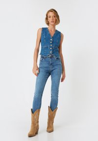 Chaleco de mezclilla con escote en V, cinco botones y dos bolsillos frontales, combinado con jeans ajustados azul claro y botas de cowboy color caqui con costuras.