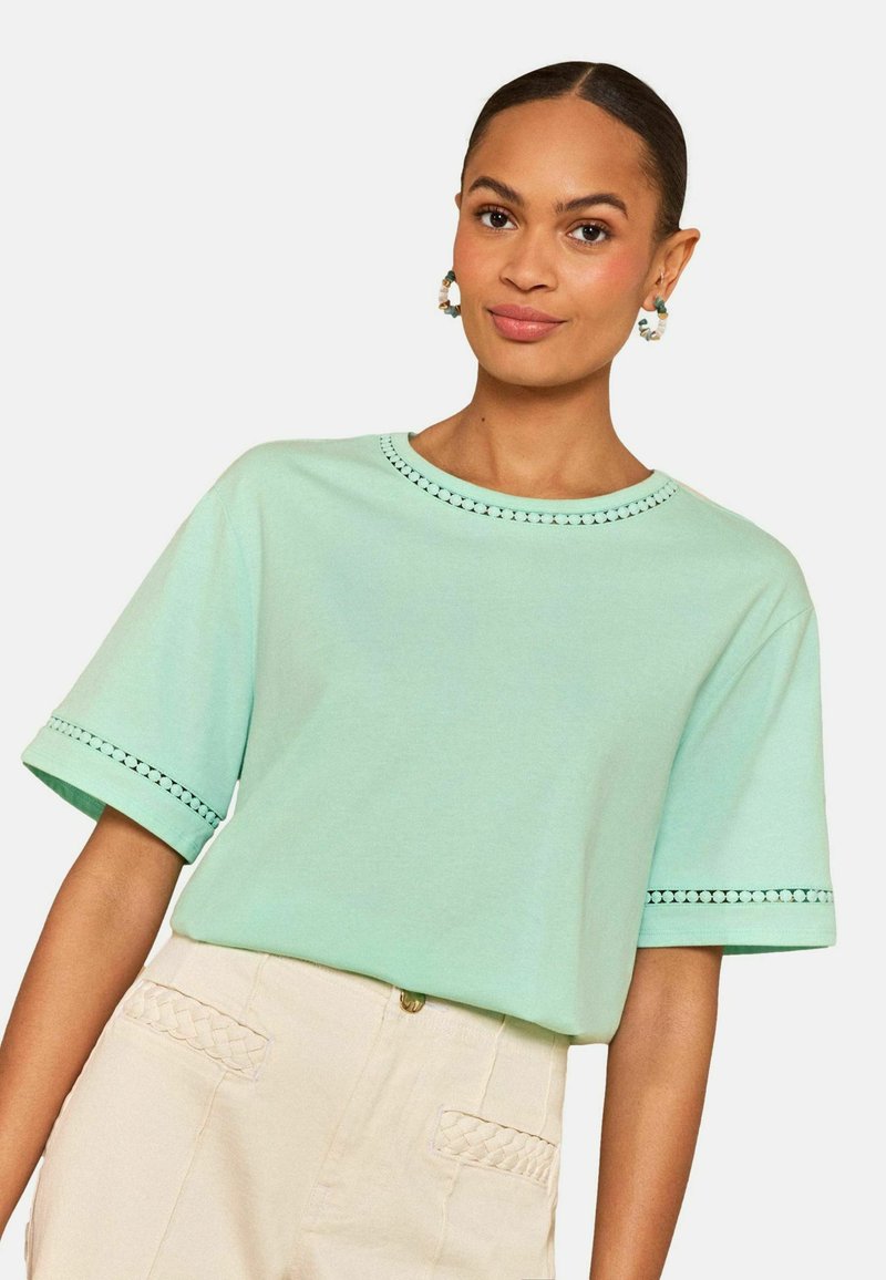 Moteris dėvi mėtų žalios spalvos trumpomis rankovėmis palaidinę su dekoratyviniu apvadu ir aukštu liemeniu kremines kelnes su pintomis kišenių detalėmis, neutraliame fone.