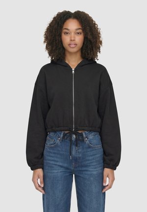 Femme aux cheveux bouclés portant un sweat à capuche court noir zippé et un jean bleu taille haute, debout devant un fond uni.