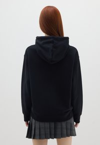 Sudadera negra vista desde atrás, con capucha, corte relajado y puños acanalados, combinada con una falda de cuadros gris plisada.