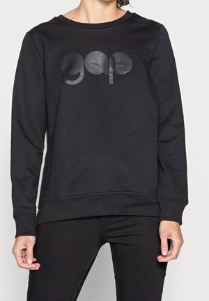 Personne portant un sweat-shirt noir à manches longues avec un logo noir brillant "gap" sur la poitrine et un pantalon noir assorti.