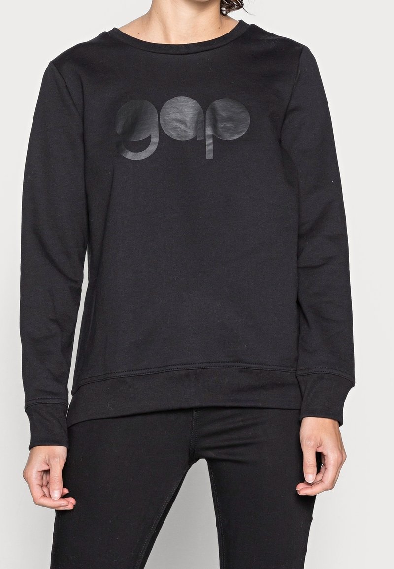 Personne portant un sweat-shirt noir à manches longues avec un logo noir brillant "gap" sur la poitrine et un pantalon noir assorti.