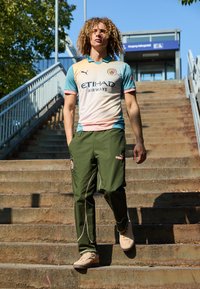 Sporttrikot in Hellblau und Creme mit pinken Akzenten, kombiniert mit olivgrünen Jogginghosen mit seitlichen Streifen und Logo-Patches.