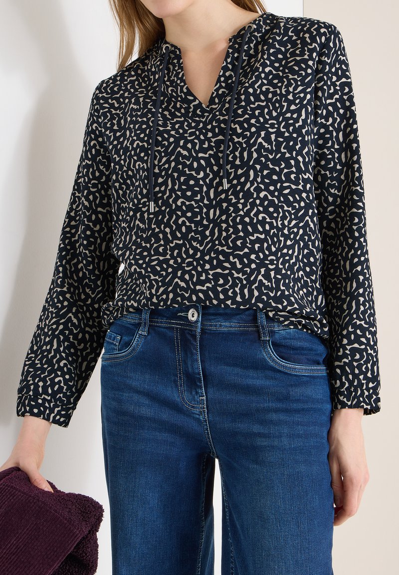 Femme portant un chemisier bleu marine à manches longues avec motifs et cordons, ainsi qu'un jean bleu, tenant un tissu violet texturé plié dans la main gauche.