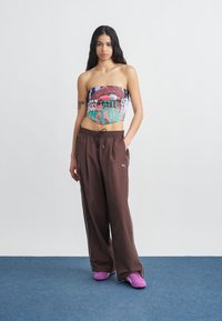 Femme aux longs cheveux foncés portant un haut coloré sans bretelles, un pantalon de survêtement marron ample Puma et des baskets violettes, debout sur un tapis bleu.