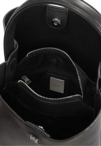 Intérieur de sac à main en cuir noir avec compartiment zippé et étiquette de marque métallique indiquant "Kazar." Fermeture à pression magnétique visible en haut.