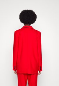 Blazer rojo con un diseño estructurado, frontal de un solo botón, solapas a pico y puños abotonados. Tejido suave con un ajuste entallado en la parte posterior.