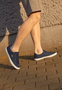Personne marchant sur une surface pavée portant des baskets bleu marine avec des semelles blanches et un short gris devant un mur texturé.