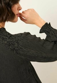 Schwarze Bluse mit weißen Punkten, mit einem rüschierten Ausschnitt und Ärmeln. Durchscheinender Stoff mit strukturierten Ärmeln.