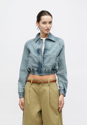 Veste en denim raccourcie bleu clair avec une fermeture éclair à l'avant, des boutons, et un ourlet élastiqué. Associée à un pantalon khaki taille haute et une ceinture marron.