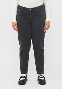 Henkilö, joka wear mustat korkeavyötäröiset farkut, valkoinen pitkähiha, valkoiset sukat ja mustat Mary Jane -tyyliset paksupohjaiset kengät, seisoo valkoista taustaa vasten.