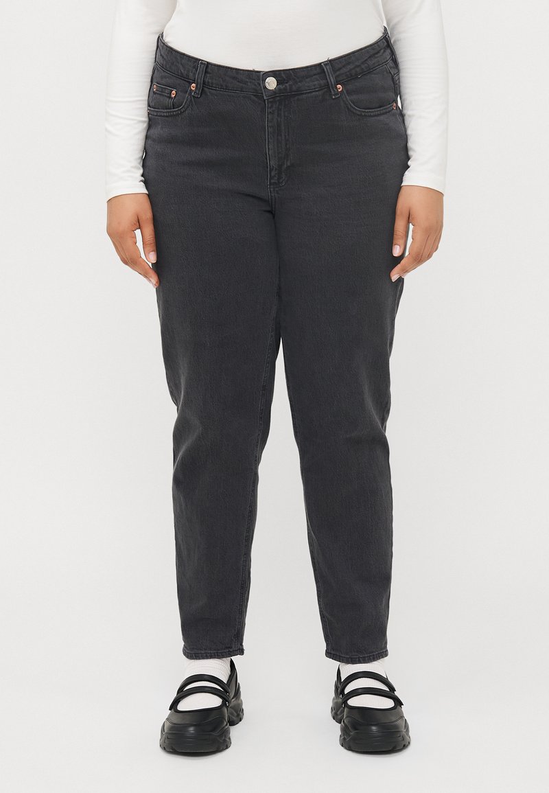 Persona vestida con jeans negros de tiro alto, blusa blanca de manga larga, calcetines blancos y zapatos negros estilo Mary Jane de suela gruesa, de pie frente a un fondo blanco.