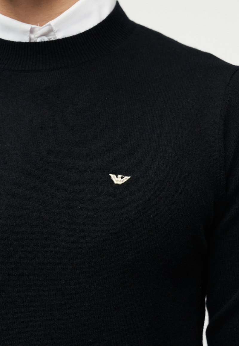 Pull en tricot noir avec un col rond côtelé, une texture douce et un petit logo aigle doré sur la poitrine gauche. Design classique, style polyvalent.