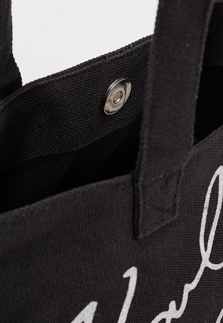 Sac fourre-tout en toile noire avec lettres argentées, dotée d'une fermeture à pression et de poignées robustes. L'intérieur présente un tissu texturé.