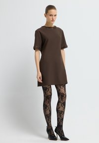 Brązowa sukienka na krótki rękaw z koralikowym dekoltem, w połączeniu z czarnymi pończochami w kwiatowy wzór i czarnymi szpilkami z ostro zakończonym noskiem. Gładki materiał, minimalistyczny design.