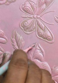 Geïllustreerd roze oppervlak met delicate motieven van vlinders en bladeren. Een hand houdt een kleine penseel en voegt details toe aan de verhoogde patronen.